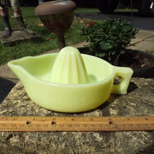 May include: A pale yellow glass lemon squeezer with a fluted center and a handle. The squeezer is sitting on a stone surface.