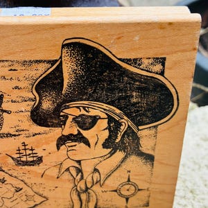 May include: Wooden stamp featuring a black illustration of a pirate with an eye patch, hat, and mustache. The design includes a map, compass, and ship. The stamp is made in the USA.