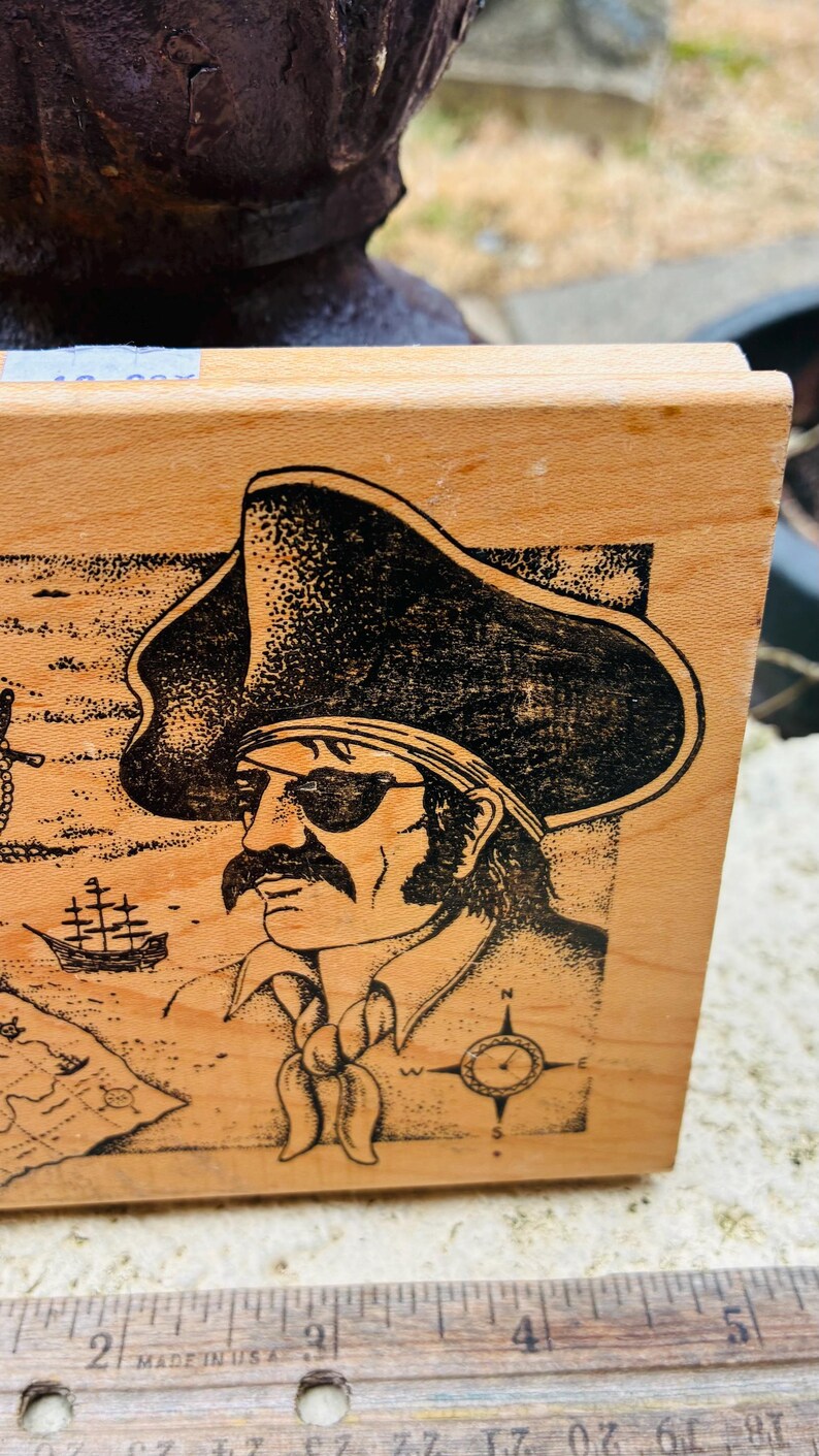 May include: Wooden stamp featuring a black illustration of a pirate with an eye patch, hat, and mustache. The design includes a map, compass, and ship. The stamp is made in the USA.