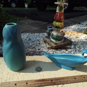 May include: Two teal blue ceramic vases, one tall and slender, the other a shallow dish with a curved handle. The vases are sitting on a gravel surface.