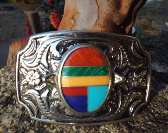 May include: A silver belt buckle with an oval inlay of red, green, yellow, blue, and turquoise stones. The buckle has a floral design.