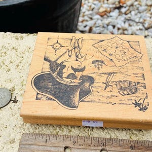 May include: Wooden stamp featuring a pirate design with a compass, treasure map, and treasure chest. The stamp is rectangular and made of light-colored wood. A ruler is placed next to the stamp, with the text "MADE IN USA".