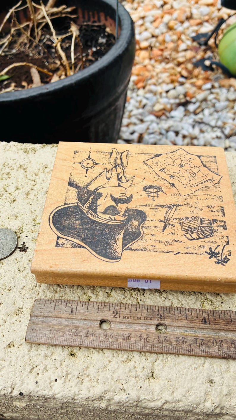 May include: Wooden stamp featuring a pirate design with a compass, treasure map, and treasure chest. The stamp is rectangular and made of light-colored wood. A ruler is placed next to the stamp, with the text "MADE IN USA".