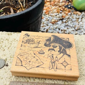 May include: Wooden stamp featuring a pirate with a map, treasure chest, and ship. The pirate wears a hat and bandana. The stamp is rectangular and sits next to a wooden ruler with inch measurements.