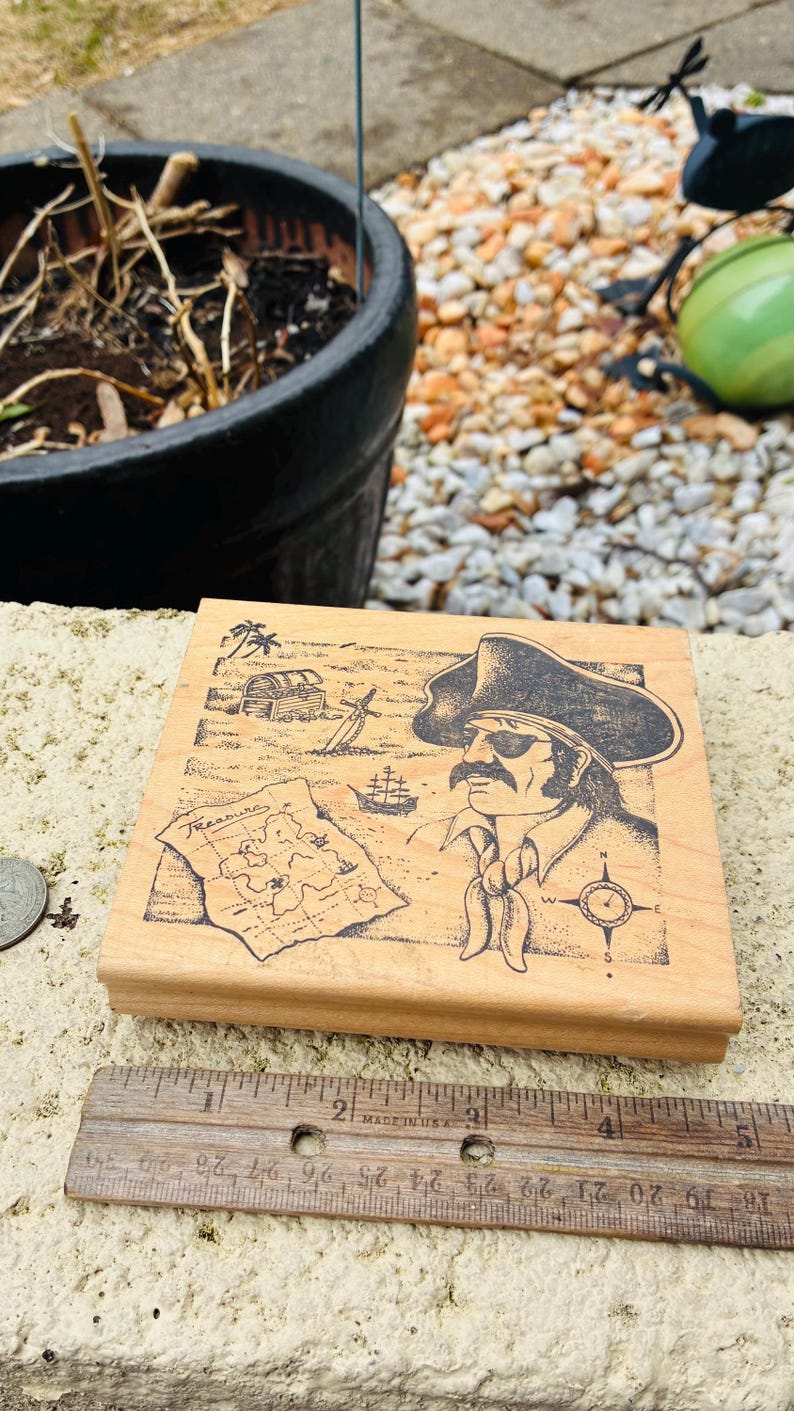 May include: Wooden stamp featuring a pirate with a map, treasure chest, and ship. The pirate wears a hat and bandana. The stamp is rectangular and sits next to a wooden ruler with inch measurements.