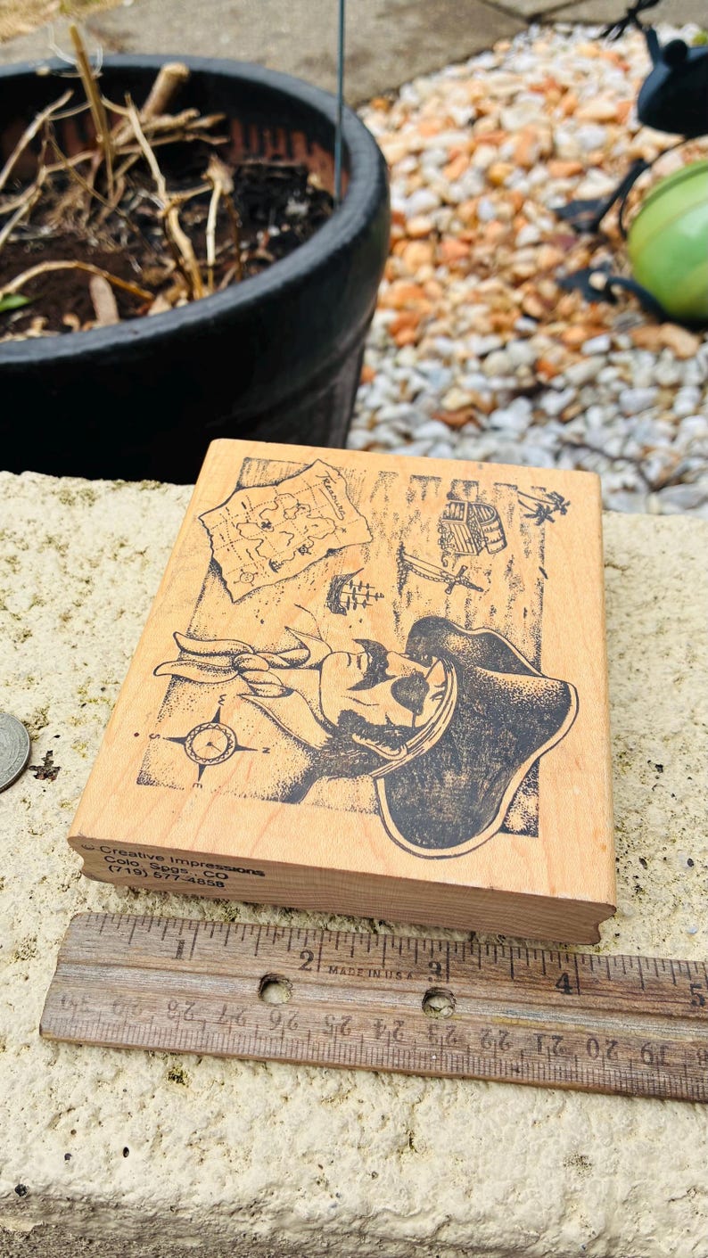 May include: Wooden stamp featuring a pirate design with a map, compass, and ship illustrations. The stamp measures approximately 3 inches in length and is made in the USA. The stamp is a craft supply.