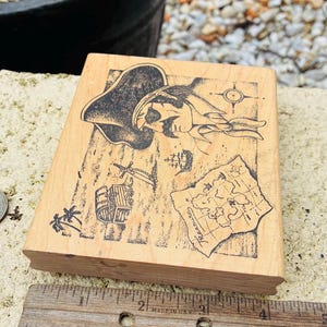 May include: A wooden stamp featuring a pirate-themed design. The stamp includes illustrations of a pirate, a treasure map, a ship, and other pirate-related elements. A wooden ruler is placed next to the stamp.