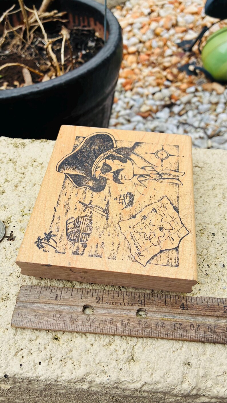May include: A wooden stamp featuring a pirate-themed design. The stamp includes illustrations of a pirate, a treasure map, a ship, and other pirate-related elements. A wooden ruler is placed next to the stamp.
