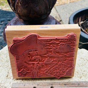 May include: A rectangular wooden stamp with a detailed red rubber design. The stamp features a cowboy with a hat, a treasure chest, and other western-themed elements. The stamp is approximately 3 inches long.