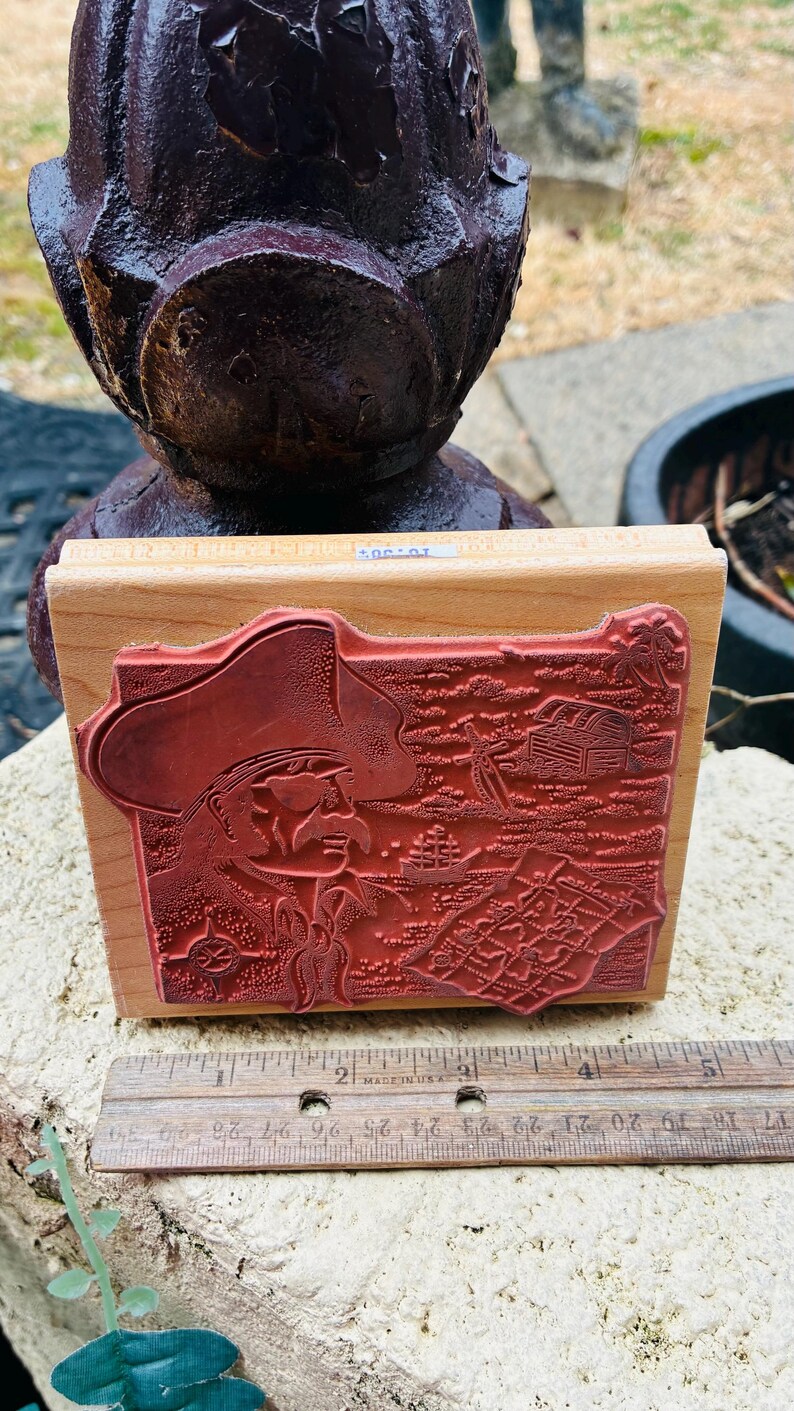 May include: A rectangular wooden stamp with a detailed red rubber design. The stamp features a cowboy with a hat, a treasure chest, and other western-themed elements. The stamp is approximately 3 inches long.