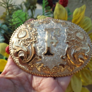 May include: A gold and silver belt buckle with a large letter F in the center. The buckle is decorated with floral designs.
