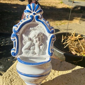 May include: A white and blue ceramic wall-mounted holy water font. The font features a relief of two cherubs and is trimmed with blue accents. The top has a decorative blue and white design, and the bottom is a rounded, shell-like shape.