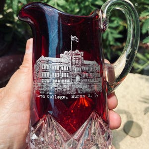 May include: A ruby red glass pitcher featuring a clear glass handle and a base with a diamond-cut pattern. The front displays an etched illustration of a building with a flag on top and the text "Huron College, Huron S. D." printed in white.