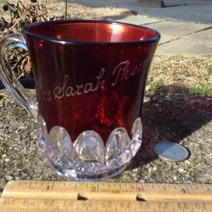 May include: A red glass mug with a clear glass base and handle. The mug has a white inscription that reads "Mrs. Sarah Thorn".
