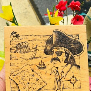 May include: Wooden stamp featuring a pirate with an eye patch, a treasure map, a treasure chest, and a compass. The word "Treasure" is written on the map. The stamp has a light brown color and is surrounded by flowers.