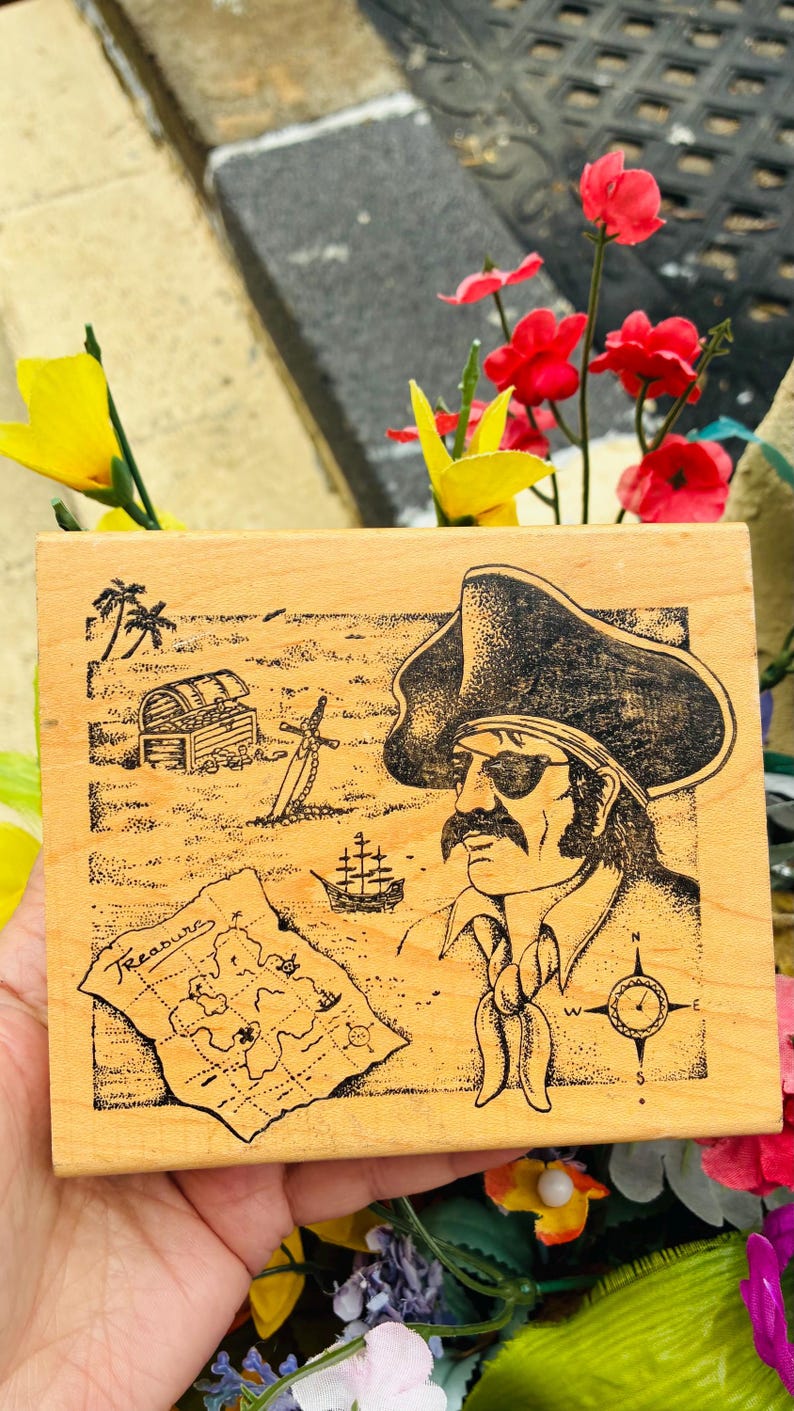 May include: Wooden stamp featuring a pirate with an eye patch, a treasure map, a treasure chest, and a compass. The word "Treasure" is written on the map. The stamp has a light brown color and is surrounded by flowers.