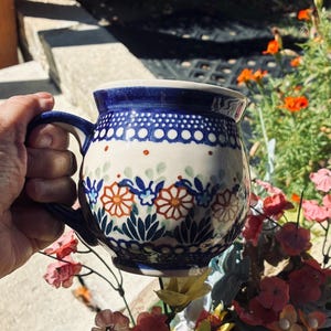 May include: A ceramic mug featuring a rounded shape and a blue and white glaze. The surface is decorated with a repeating pattern of orange flowers, blue leaves, and small dots. The handle is curved and matches the blue rim and base accents.