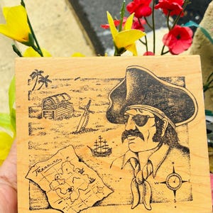 May include: Wooden block featuring a pirate illustration. The design includes a treasure map with the word "Treasure", a pirate with an eye patch, a treasure chest, a sword, and a ship. The artwork is in black ink on a light wood background.