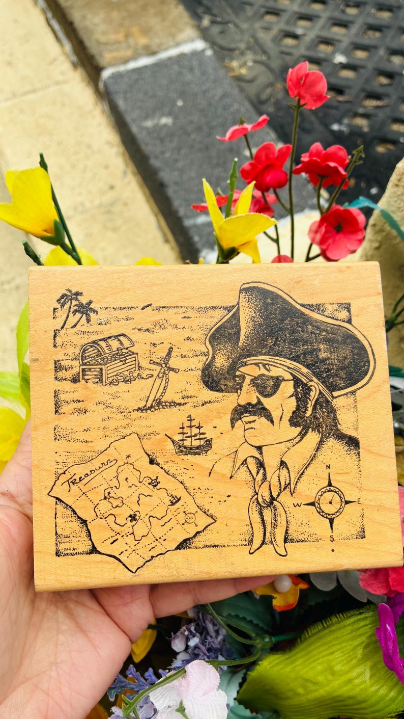 May include: Wooden block featuring a pirate illustration. The design includes a treasure map with the word "Treasure", a pirate with an eye patch, a treasure chest, a sword, and a ship. The artwork is in black ink on a light wood background.