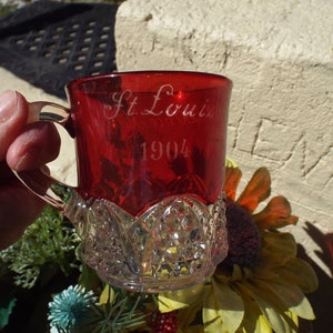 May include: A red glass cup with a clear glass handle and base. The cup is etched with the words "St. Louis 1904".