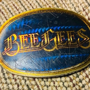 May include: Oval belt buckle with a blue background and the word "BEE GEES" in gold lettering. The buckle has a gold border and a textured surface, showing signs of wear.