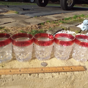 May include: Set of five clear glass tumblers with a red rim. The glasses have a diamond pattern and are arranged in a row.