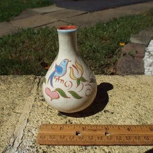 May include: A small, off-white ceramic vase with a crackle glaze. It features hand-painted designs of a blue bird, pink hearts, and yellow flowers with green leaves. The rim is red with a light blue band. A wooden ruler is next to the vase.