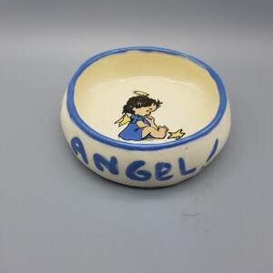 May include: A small, off-white ceramic bowl with a blue rim and the word "ANGEL" in blue lettering. The interior features a cartoon angel in a blue dress and a gold star. The bowl is round and sits on a gray surface.