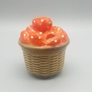 May include: A ceramic lidded jar shaped like a woven brown basket filled with bright red strawberries. The lid features a cluster of sculpted strawberries with small white dots, while the base mimics the texture of a woven wicker basket.