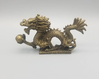 May include: A gold-colored brass dragon figurine holding a pearl in its claws. The dragon is curled in a defensive position with its tail curled around its body. The figurine is detailed with scales and intricate designs.