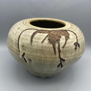 Puede incluir: Un jarrón de cerámica blanco con un diseño floral marrón. El jarrón tiene una forma redondeada y una abertura ancha.