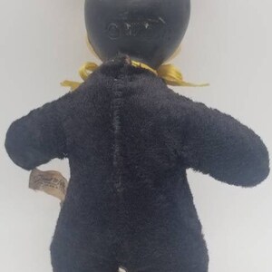 May include: A black plush Mickey Mouse doll with yellow fabric ears and a yellow bow tie. The doll is wearing a black felt suit and has a black felt head. The doll is standing on a white surface. The doll has a tag on its leg that says "Disney".