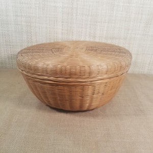 May include: A round, lidded basket made of woven light brown wicker. The basket has a rounded base and a matching lid that fits snugly on top. The weave pattern is consistent throughout, creating a textured surface.