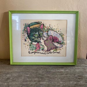May include: A framed print featuring two cats, one green and one pink, in a loving embrace. The green-eyed cat wears a hat and bow tie. The print is in a green frame and includes the text "everyone needs to be loved."