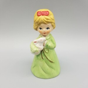 Puede incluir: Figura de cerámica de una niña con un vestido verde claro, sosteniendo un cordero blanco. La niña tiene cabello rubio con un lazo rojo y ojos azules. La figura es un objeto decorativo.