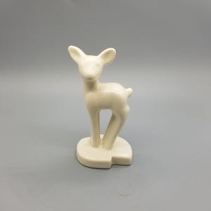 以下が含まれることがあります： クリーム色のセラミック製の鹿の置物。鹿は、耳を立てて小さな長方形の台座の上に立っています。置物は滑らかで光沢のある仕上がりで、灰色の背景に配置されています。鹿の高さは約13cmです。
