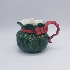 Puede incluir: Una jarra de crema de cerámica verde con forma de saco de Navidad con una cinta y un lazo rojos. La jarra tiene un interior blanco y un asa roja.