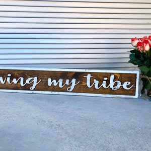 May include: A rectangular wooden sign with the phrase "loving my tribe" in white, raised lettering. The sign has a dark brown center and a white border. A vase of red and white roses sits to the right.