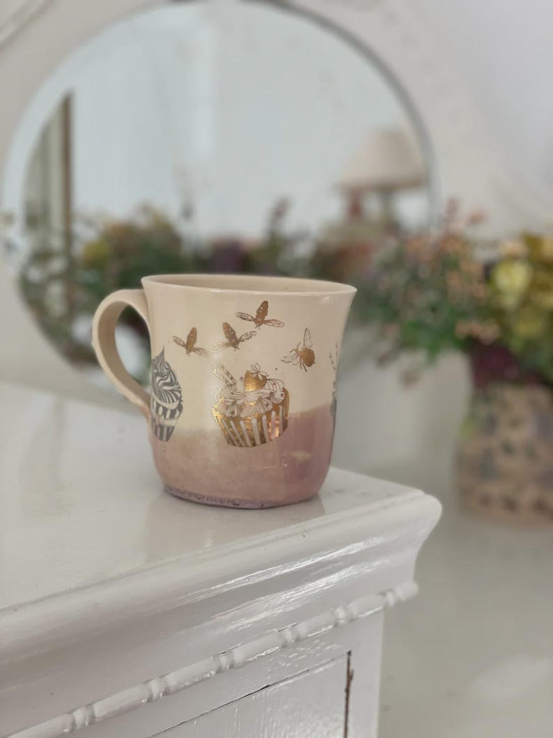 May include: A ceramic mug with a cream-colored upper section and a dusty rose-colored base. The mug features metallic gold illustrations of cupcakes and flying bees encircling the exterior, along with a curved handle on the left side.