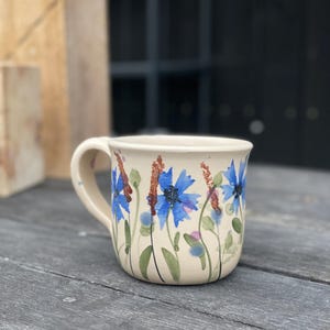 Puede incluir: Taza de cerámica color crema con diseño floral. La taza presenta acianos azules pintados a mano, tallos marrones y hojas verdes. La taza tiene un asa curva y está sobre una superficie de madera gris desgastada.