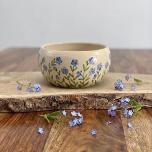 Op de afbeelding: Een ronde keramische kom met een crèmekleurige basis voorzien van een handgeschilderd patroon van kleine blauwe vergeet-mij-nietjes met groene stengels en bladeren. De kom heeft een gladde, gebogen rand en een glanzende afwerking.