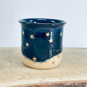 May include: A ceramic mug featuring a two-tone design with a deep navy blue upper section and a cream-colored base. The exterior is decorated with small, gold-colored star motifs scattered across the surface, creating a celestial pattern.