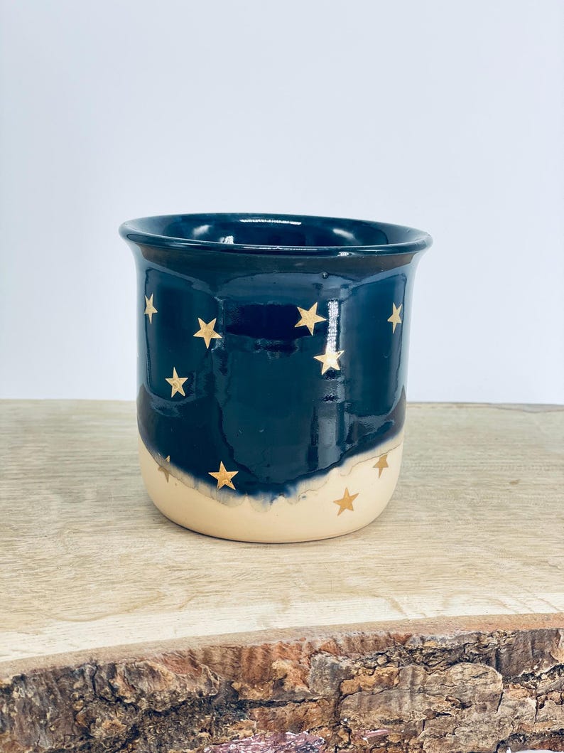 May include: A ceramic mug featuring a two-tone design with a deep navy blue upper section and a cream-colored base. The exterior is decorated with small, gold-colored star motifs scattered across the surface, creating a celestial pattern.