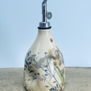 May include: A cream-colored ceramic oil dispenser with a tapered body and a silver-toned metal pour spout. The surface features a botanical print with blue and green floral illustrations, including delicate stems, leaves, and umbel-shaped flowers.