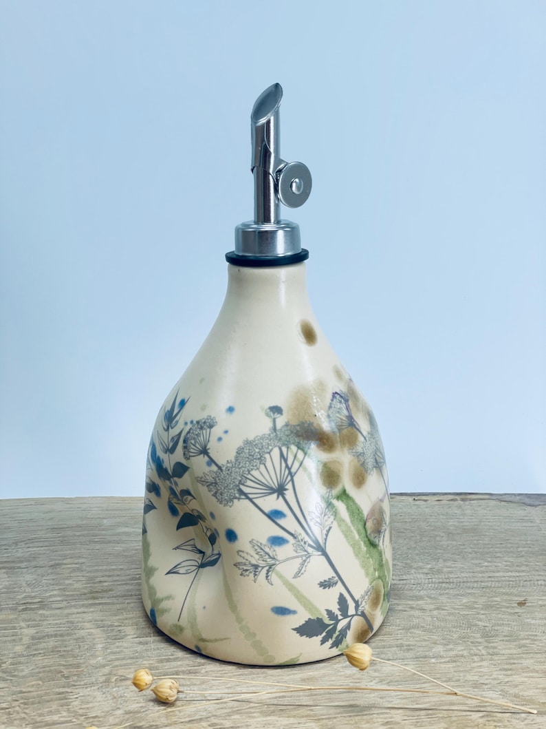 May include: A cream-colored ceramic oil dispenser with a tapered body and a silver-toned metal pour spout. The surface features a botanical print with blue and green floral illustrations, including delicate stems, leaves, and umbel-shaped flowers.