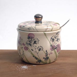 May include: A cream-colored ceramic sugar bowl with a matching lid and a small metal spoon. The exterior features a botanical pattern of black line-drawn wildflowers and soft purple watercolor-style circular accents on the body and lid.