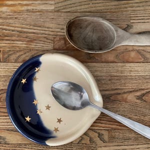 May include: A round ceramic spoon rest featuring a two-tone design with a deep navy blue section and a cream-colored section. The surface is decorated with small, metallic gold-colored star shapes scattered across both colors. A silver metal spoon rests inside.