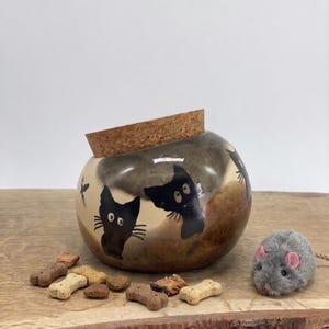 Pode incluir: Um recipiente redondo de cabaça na cor castanho-claro com tampa de cortiça, decorado com silhuetas de gatos pretos pintadas à mão. O recipiente está rodeado por vários pequenos petiscos para animais em forma de osso e um rato de brinquedo cinzento.