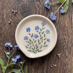Pode incluir: Um descanso de colher redondo em cerâmica branca com um design pintado à mão de flores miosótis azuis com caules e folhas verdes. O centro do prato apresenta um grupo destas pequenas flores azuis, com as bordas lisas e simples.