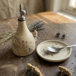 May include: A cream-colored ceramic oil dispenser with a metal pour spout and a matching small round ceramic dish. Both pieces feature a delicate botanical print of dark gray and purple wildflowers. A silver metal spoon rests on the dish.